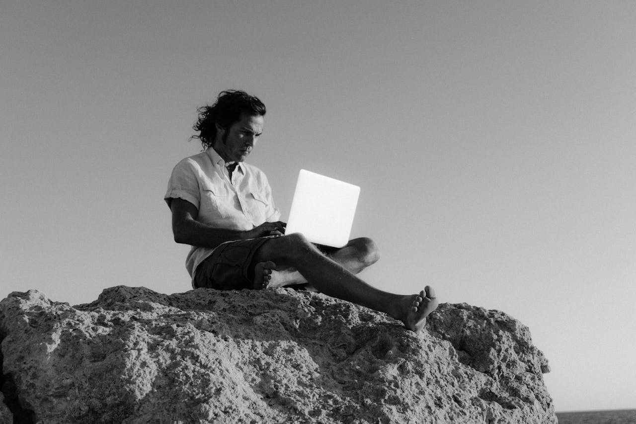A digital nomad works on a laptop while sitting on a rocky cliff, embodying remote work lifestyle.